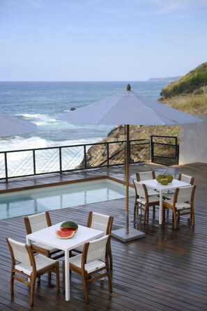 Área ao lado da piscina com mesa e cadeiras, vista para o oceano e um corrimão.