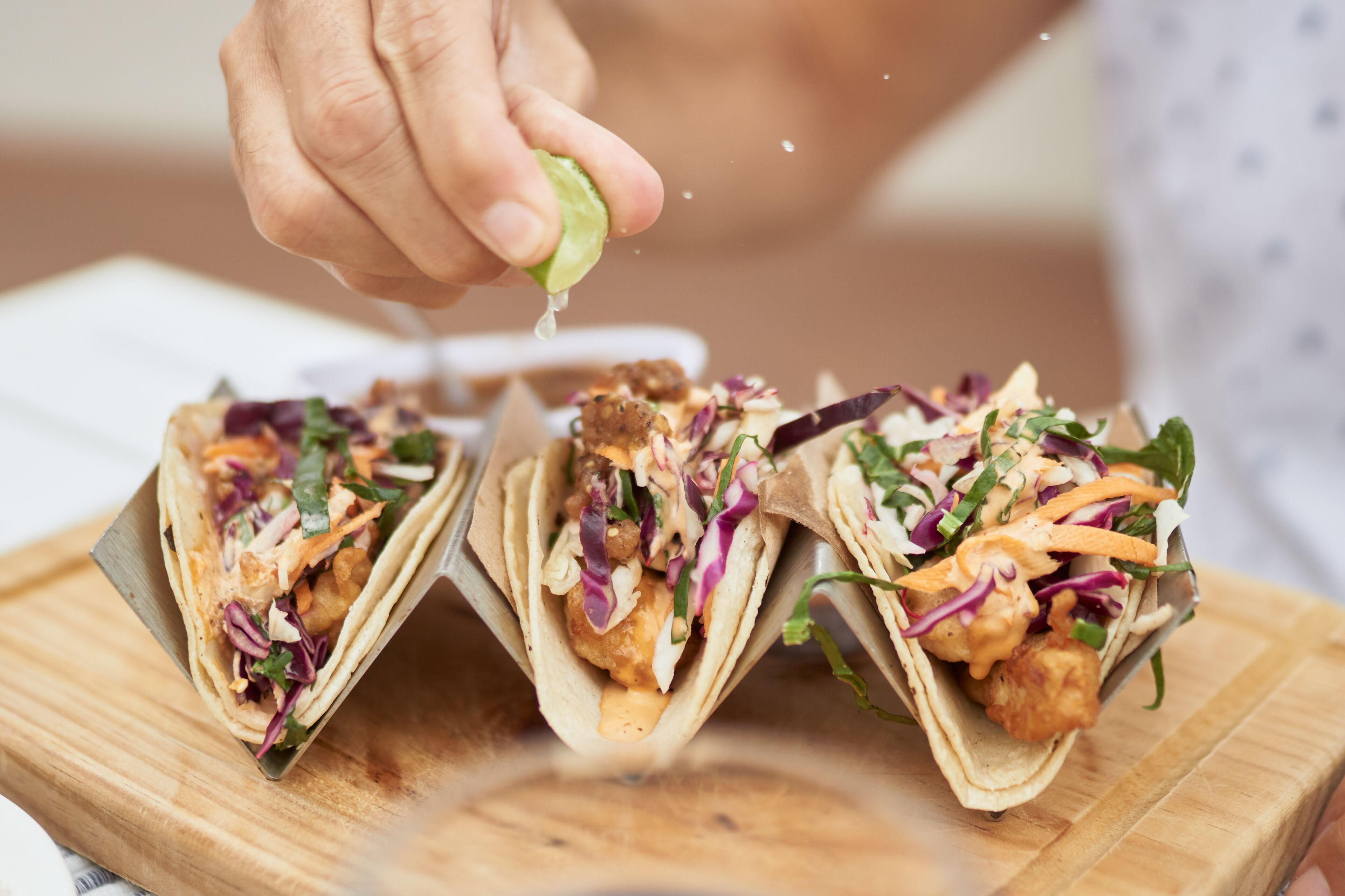 Tres tacos sobre una tabla de madera, con una mano exprimiendo un limón.
