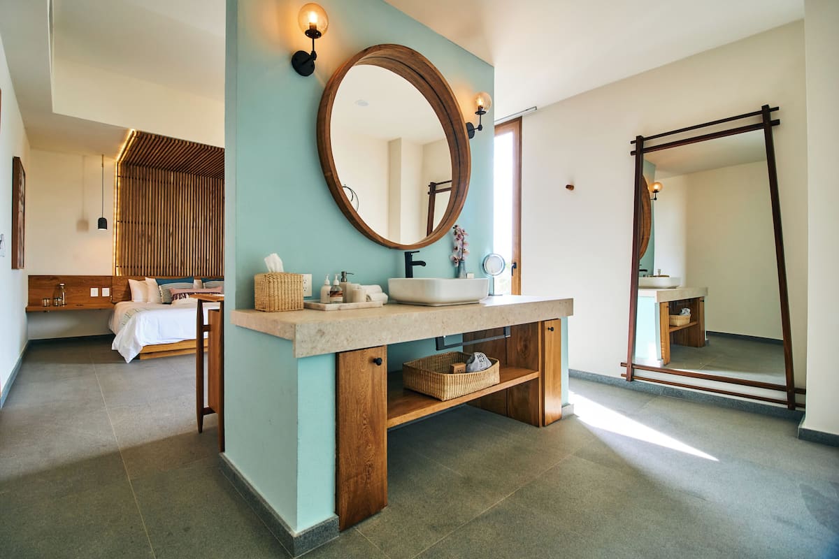 A modern bathroom with a large mirror, wooden vanity, and a bed in the adjacent room.