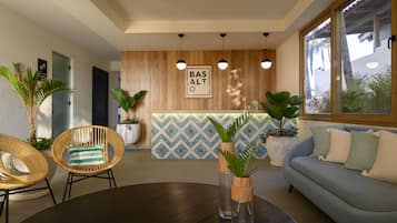Un área de recepción moderna con un mostrador de madera, dos sillas de mimbre, una mesa redonda y un sofá azul con cojines verdes.