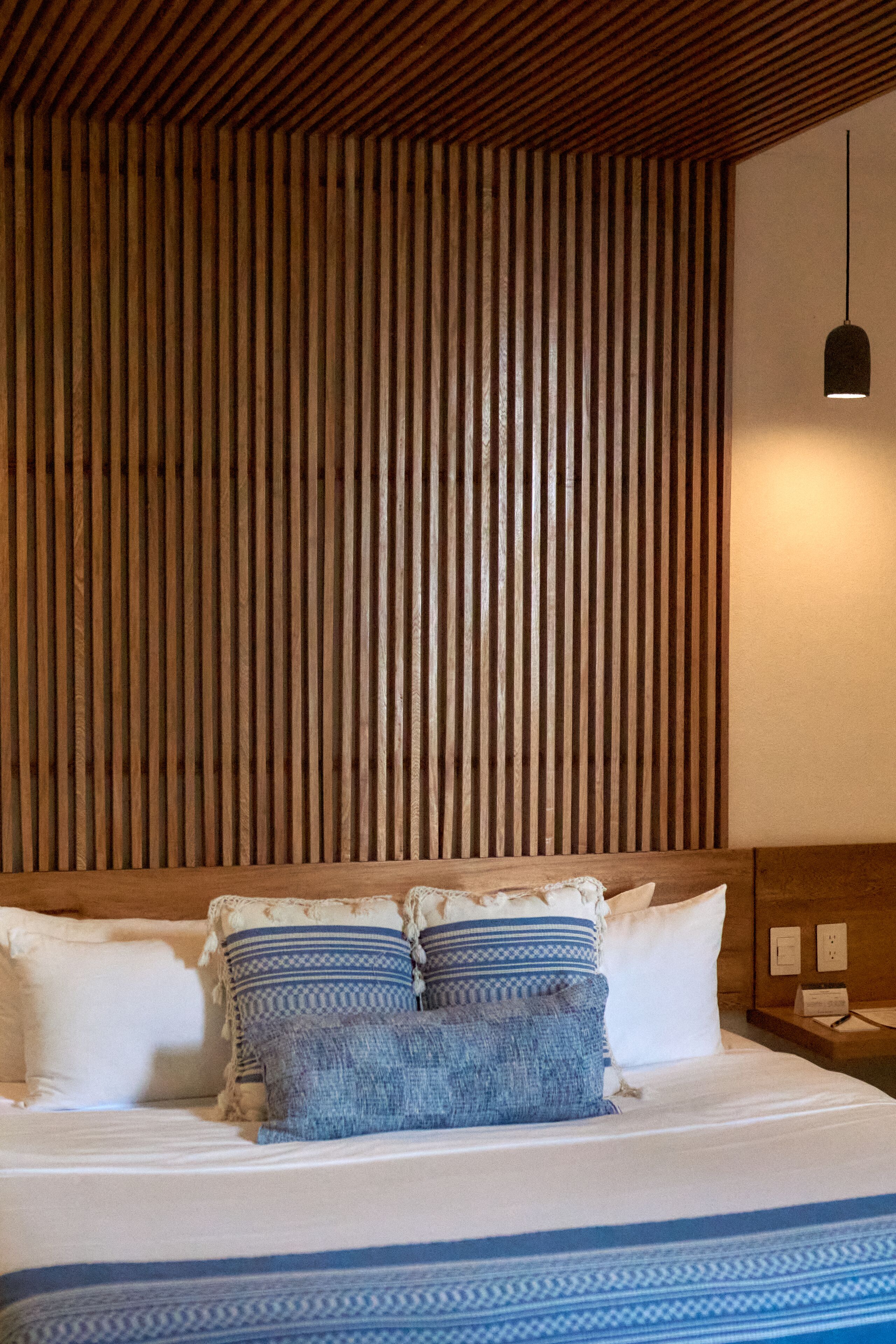 Una cama bien hecha con una colcha azul y blanca a rayas, dos almohadas con diseños azules y blancos, y un cabecero de madera con listones.