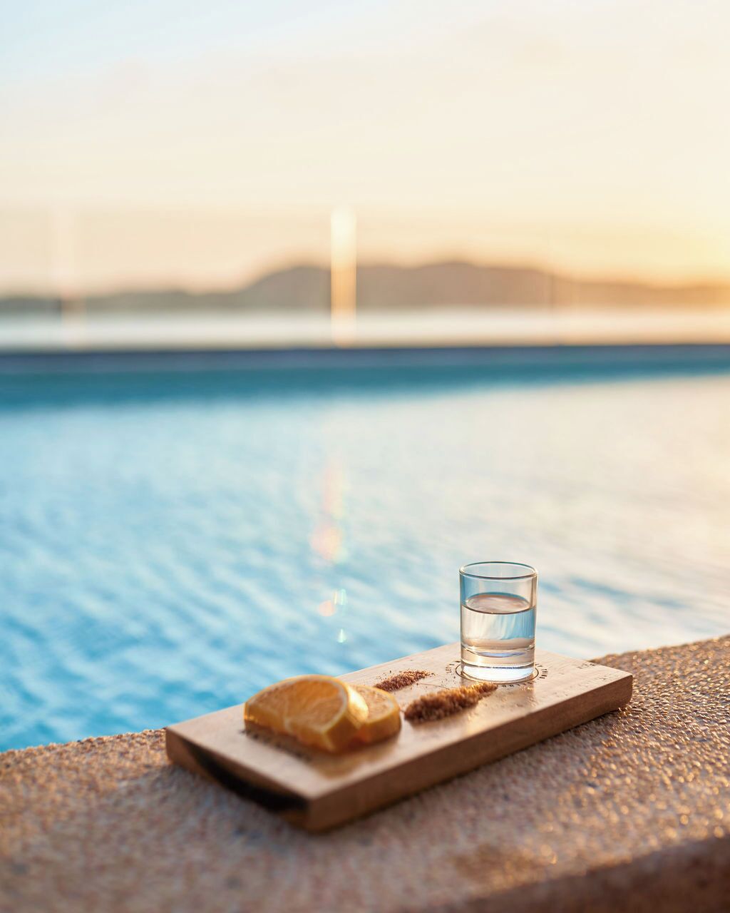 Un verre d’eau et une tranche d’orange sur une planche en bois, près d’une piscine, avec une vue lointaine.