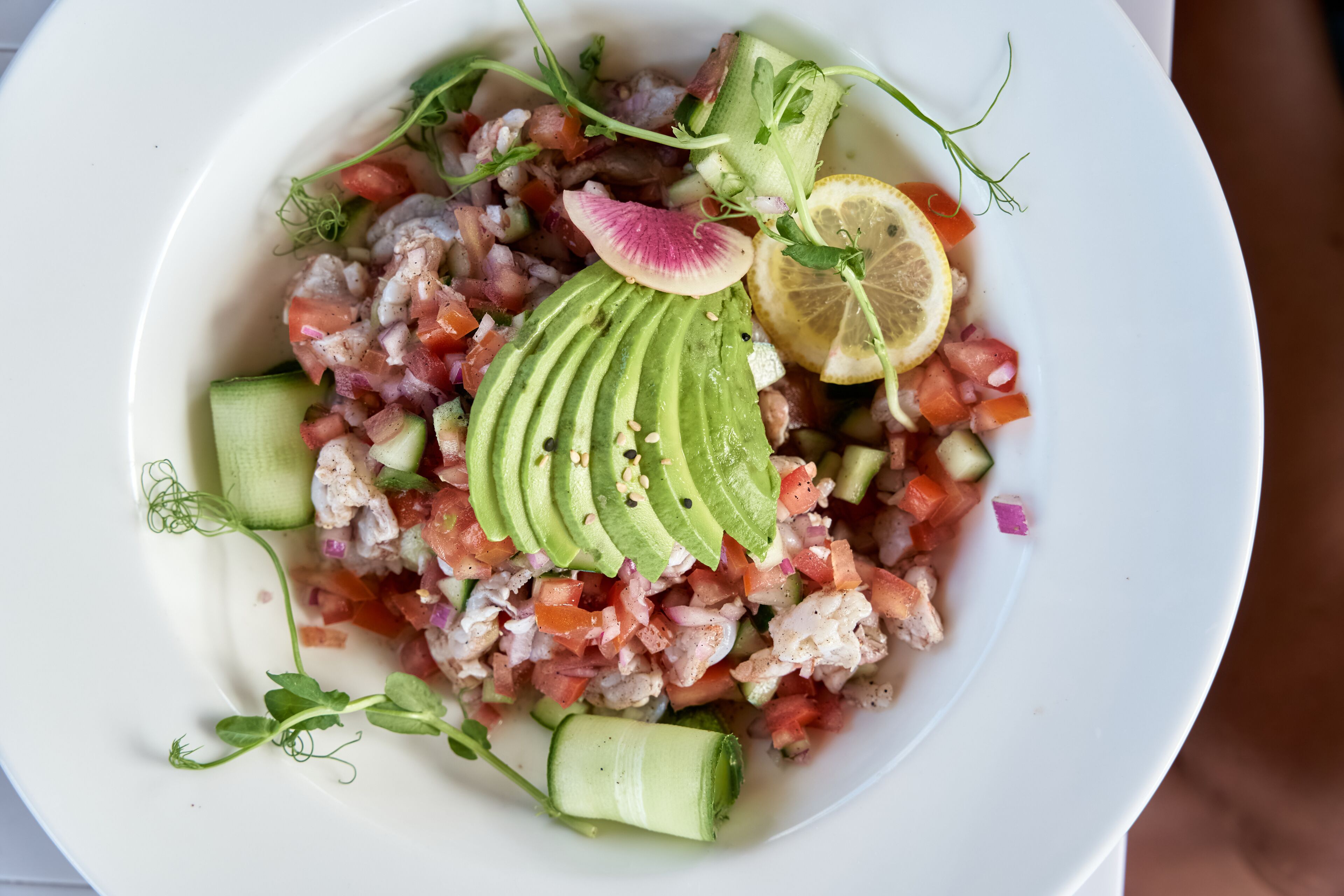 Une assiette blanche contenant une salade de fruits de mer, des tranches d’avocat, un citron et du concombre.