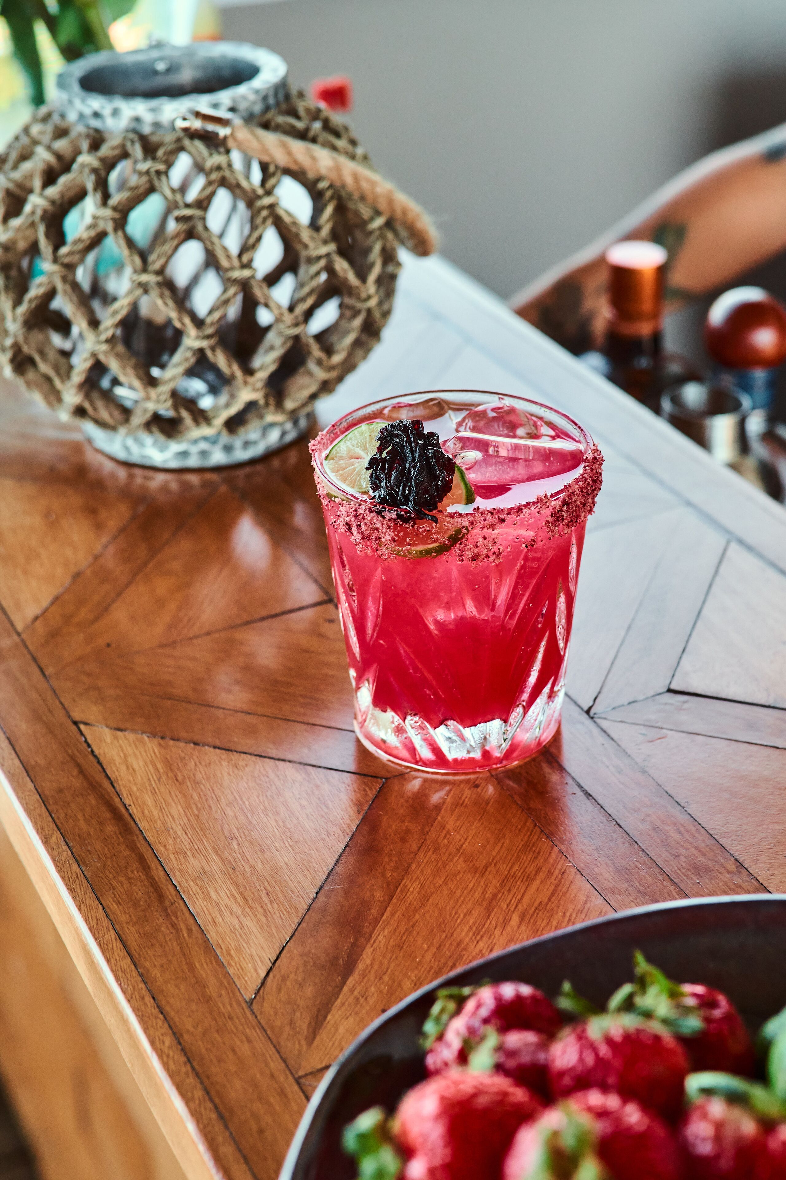 A cocktail glass with a red drink, garnished with a lime and a black berry, on a wooden table.