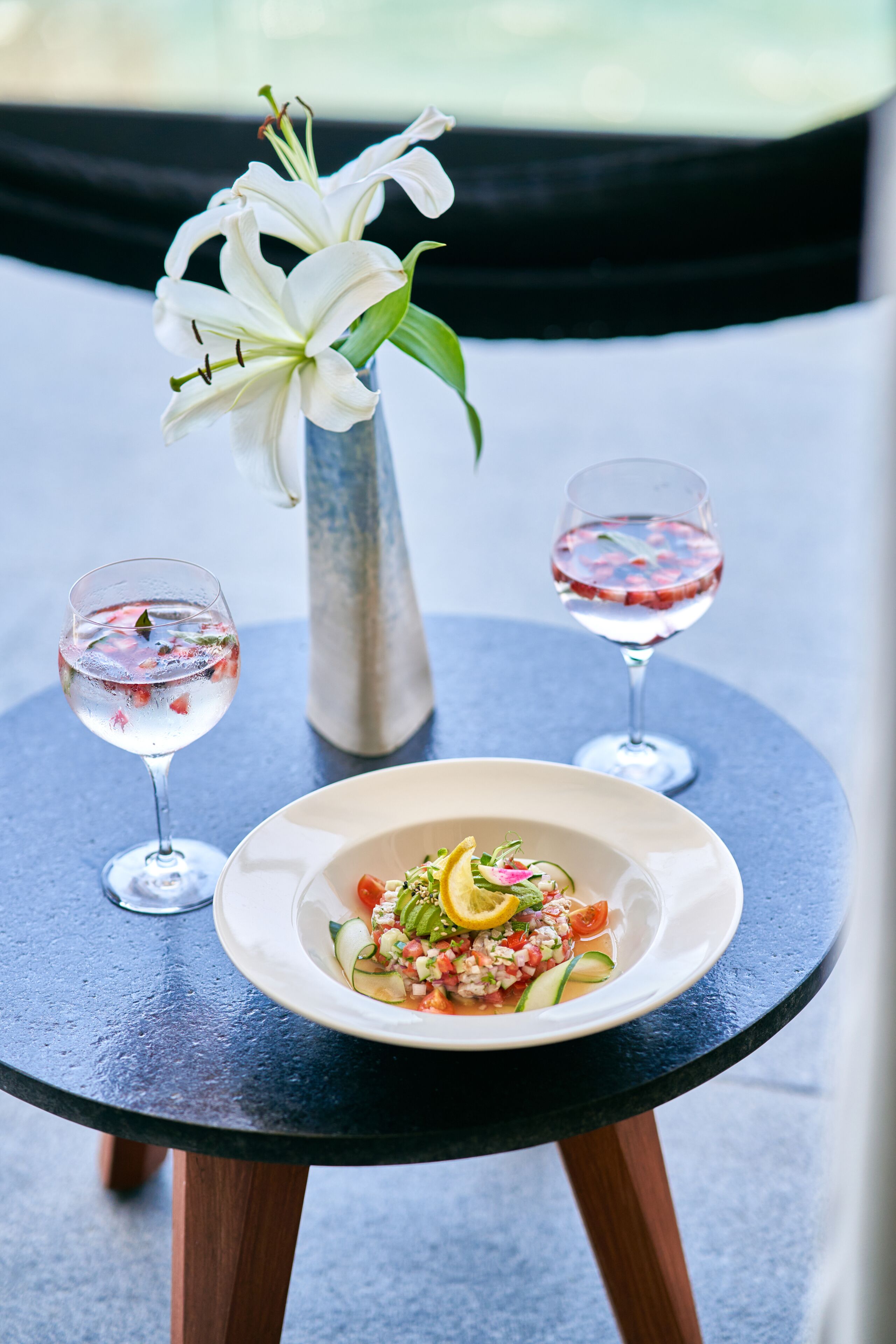 A table with a plate of food, two glasses of drink, and a vase of flowers.