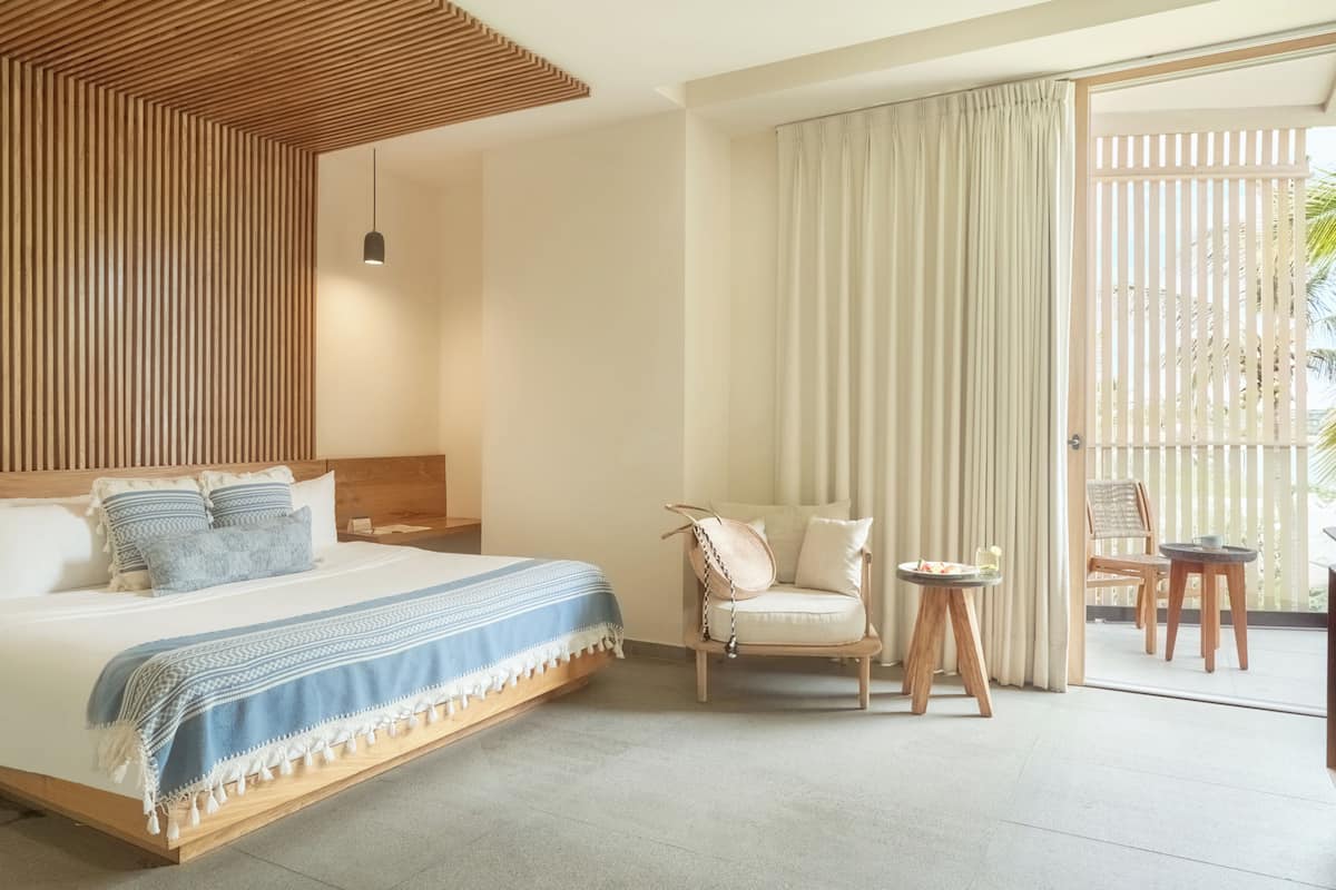 A hotel room with a large bed, a wooden ceiling, a balcony with a view, and a small table with a chair.