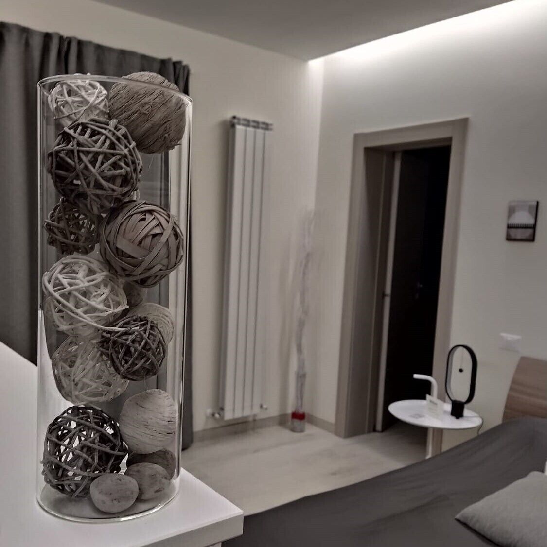 Un dormitorio moderno con un florero de vidrio lleno de esferas decorativas sobre una mesita de noche, una pequeña mesa redonda con un espejo y un radiador al fondo.