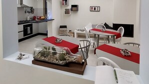 Une cuisine moderne avec des meubles blancs, un coin repas avec des chaises blanches et des nappes rouges, et une petite bibliothèque contenant des livres et des objets décoratifs.