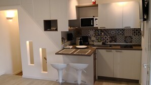 Eine kompakte Küche mit weißen Schränken, einer Mikrowelle, einem Toaster und einem Spülbecken. Zwei Hocker und eine kleine Ablagefläche befinden sich über der Arbeitsplatte.