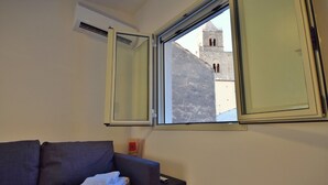Ein Zimmer mit grauem Sofa, einem Fenster mit Blick auf ein Gebäude und einer Klimaanlage an der Wand.