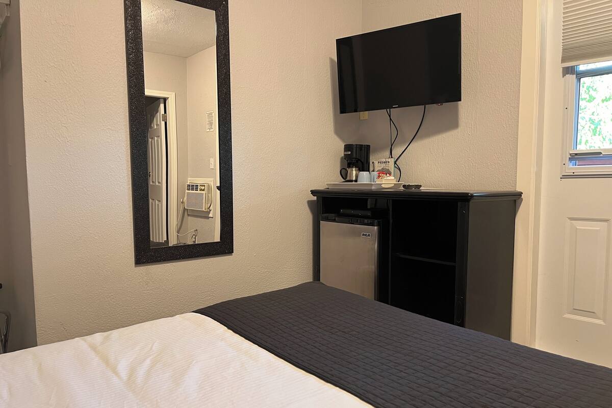 A hotel room with a bed, a mirror, a television mounted on the wall, a small desk with a coffee maker, and a window with blinds.