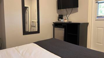 A hotel room with a bed, a mirror, a television mounted on the wall, a small desk with a coffee maker, and a window with blinds.