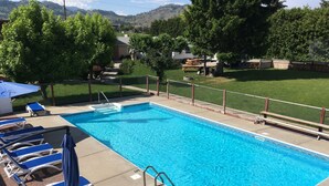 Une piscine rectangulaire à l’eau d’un bleu limpide, entourée d’une terrasse en béton. On y trouve des chaises longues avec des coussins bleus et des structures blanches, ainsi qu’un parasol bleu. L’espace piscine est délimité par une clôture en treillis métallique et, au fond, on aperçoit une terrasse en bois.