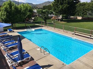 A rectangular swimming pool with clear blue water, surrounded by a concrete deck. There are lounge chairs with blue cushions and white frames, and a blue umbrella. The pool area is fenced with a chain-link fence and has a wooden deck in the background.