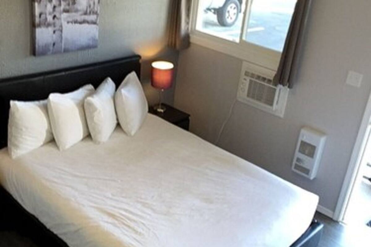 A bedroom with a bed, pillows, a nightstand with a lamp, a window with a view, and a wall-mounted air conditioning unit.