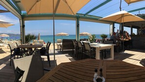 Eine Terrassengaststätte mit Holztischen und -stühlen, Sonnenschirmen und Meerblick.
