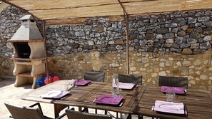 An outdoor dining area with a stone wall, a stone chimney, and a table set with purple napkins and plates.