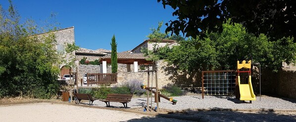 A playground with a yellow slide and a climbing structure, surrounded by a stone building with a tiled roof.