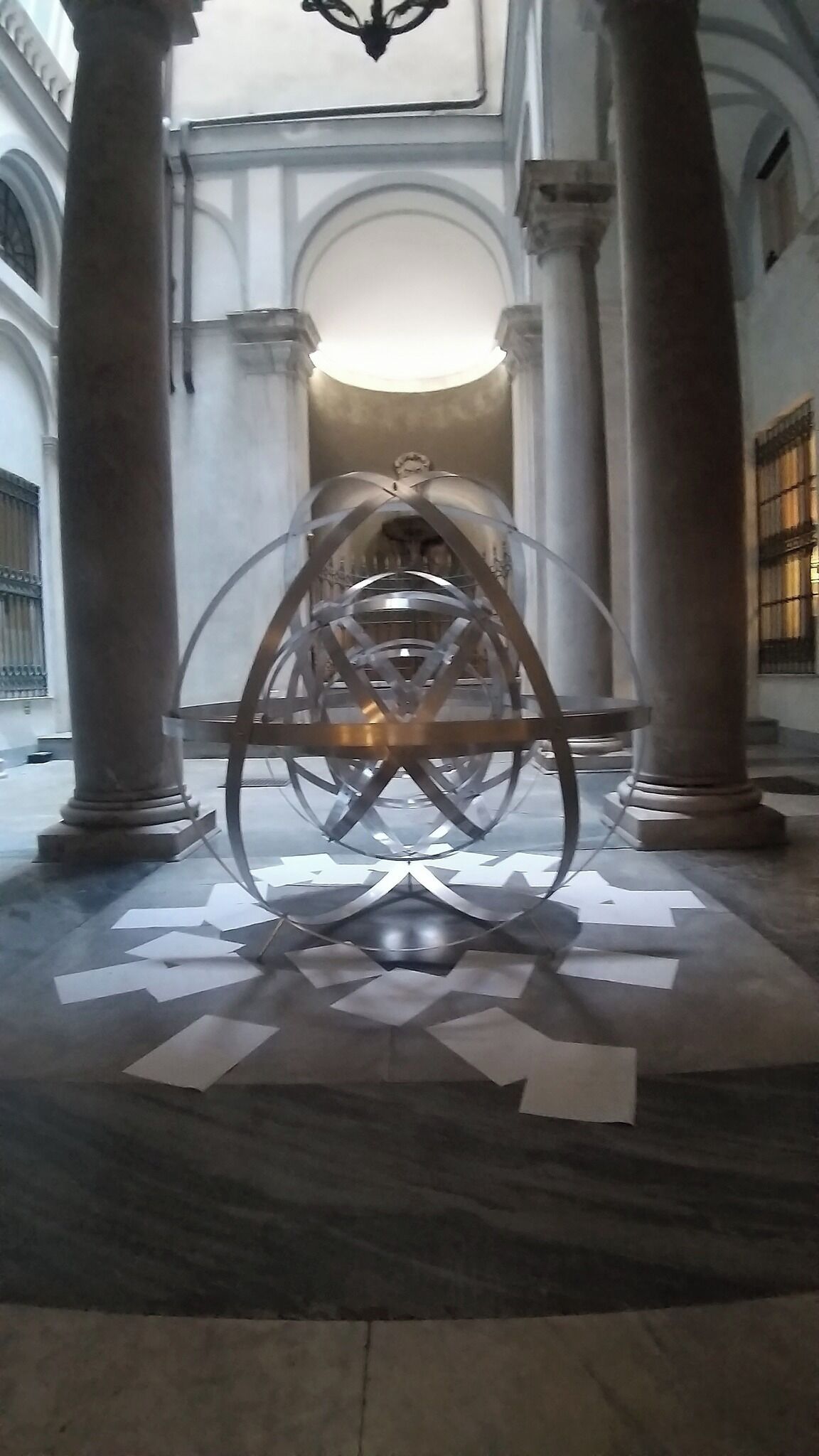 Eine große, metallische Skulptur in einem geräumigen, hell erleuchteten Atrium mit hohen Decken und klassischer Architektur, darunter Säulen und Bögen.