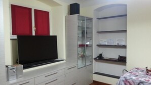 A compact room with a flat-screen TV mounted on a cabinet, a glass-fronted shelving unit, and a small table with a patterned tablecloth. - SMITHS & HERREROS (Codie CIN IT010025C2Q2M67QJW) (Genova)