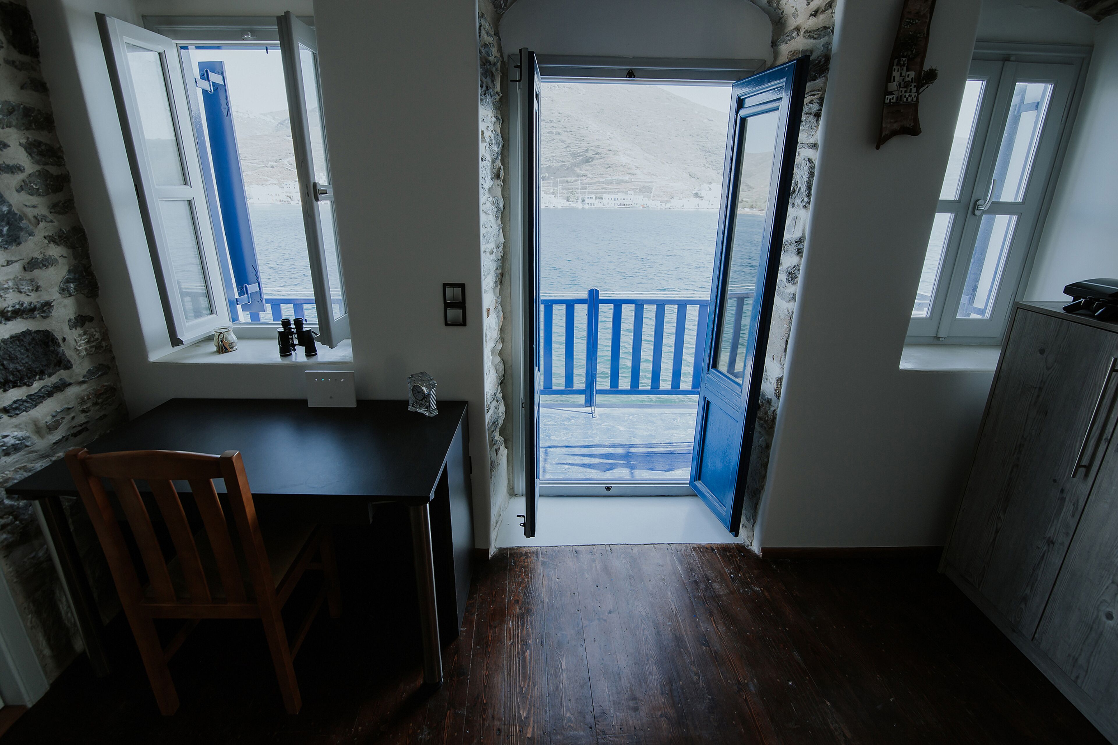 Une pièce avec un bureau en bois, une chaise et une vue sur un balcon à travers une porte ouverte, le balcon étant doté d’une balustrade bleue.