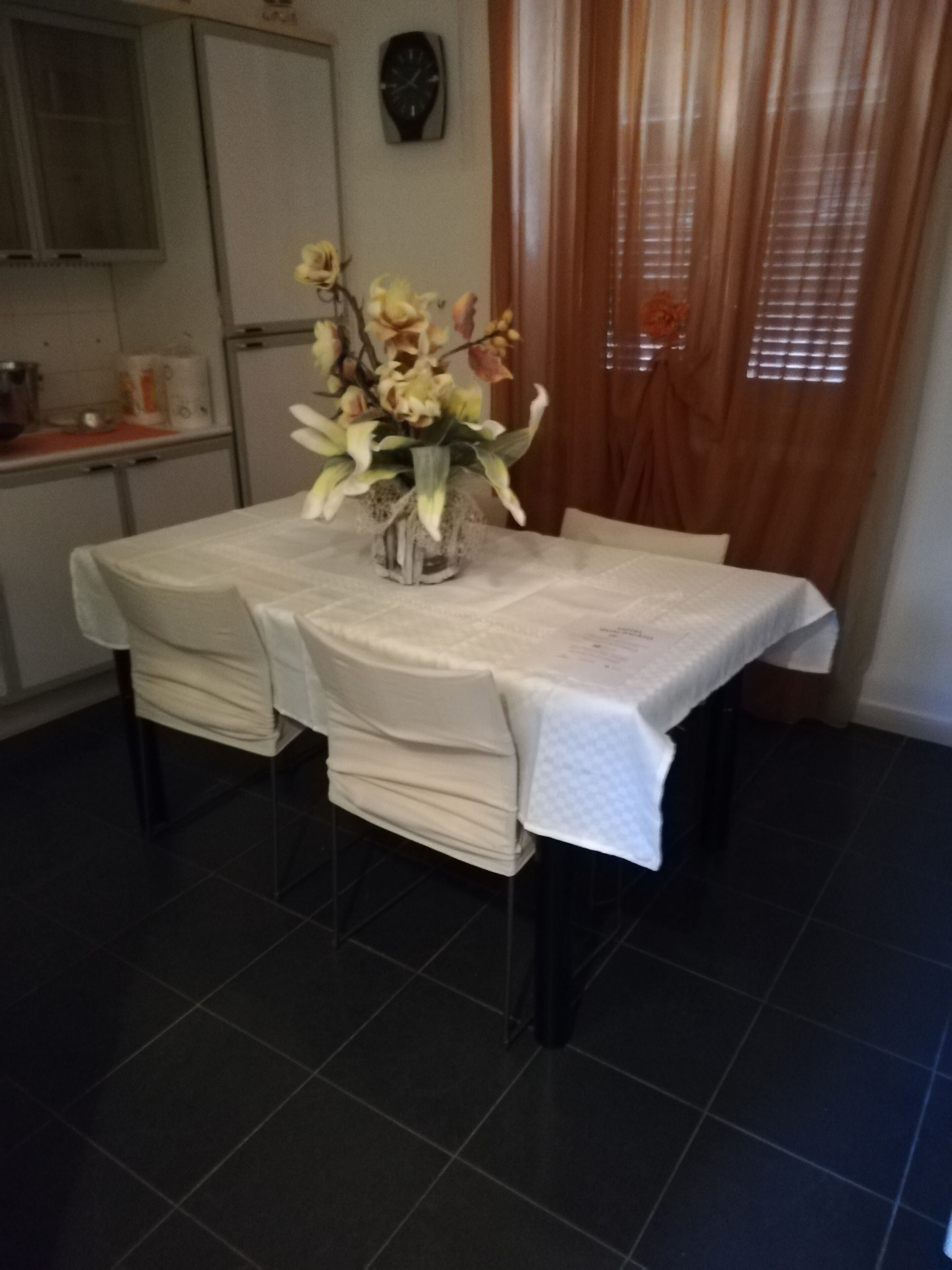 Ein Esstisch mit weißer Tischdecke, umgeben von Stühlen mit weißen Hussen. Auf dem Tisch steht eine Vase mit Blumen.