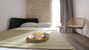 A neatly made bed with a tray of breakfast items, a wicker chair, and a stone wall in the background. - Funduq iblea hospitality - ground floor apartment (Pozzallo)