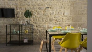 A dining area with a stone wall, a TV mounted on the wall, a glass shelf with dishes, and yellow chairs around a table set for four. - Funduq iblea hospitality - ground floor apartment (Pozzallo)