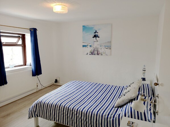 A bedroom with a bed, a window with curtains, a painting of a lighthouse, and a bedside lamp. - Cosy & Comfortable Entire Holiday Home, Llanelli. Fully equipped. Dog friendly (Llanelli)