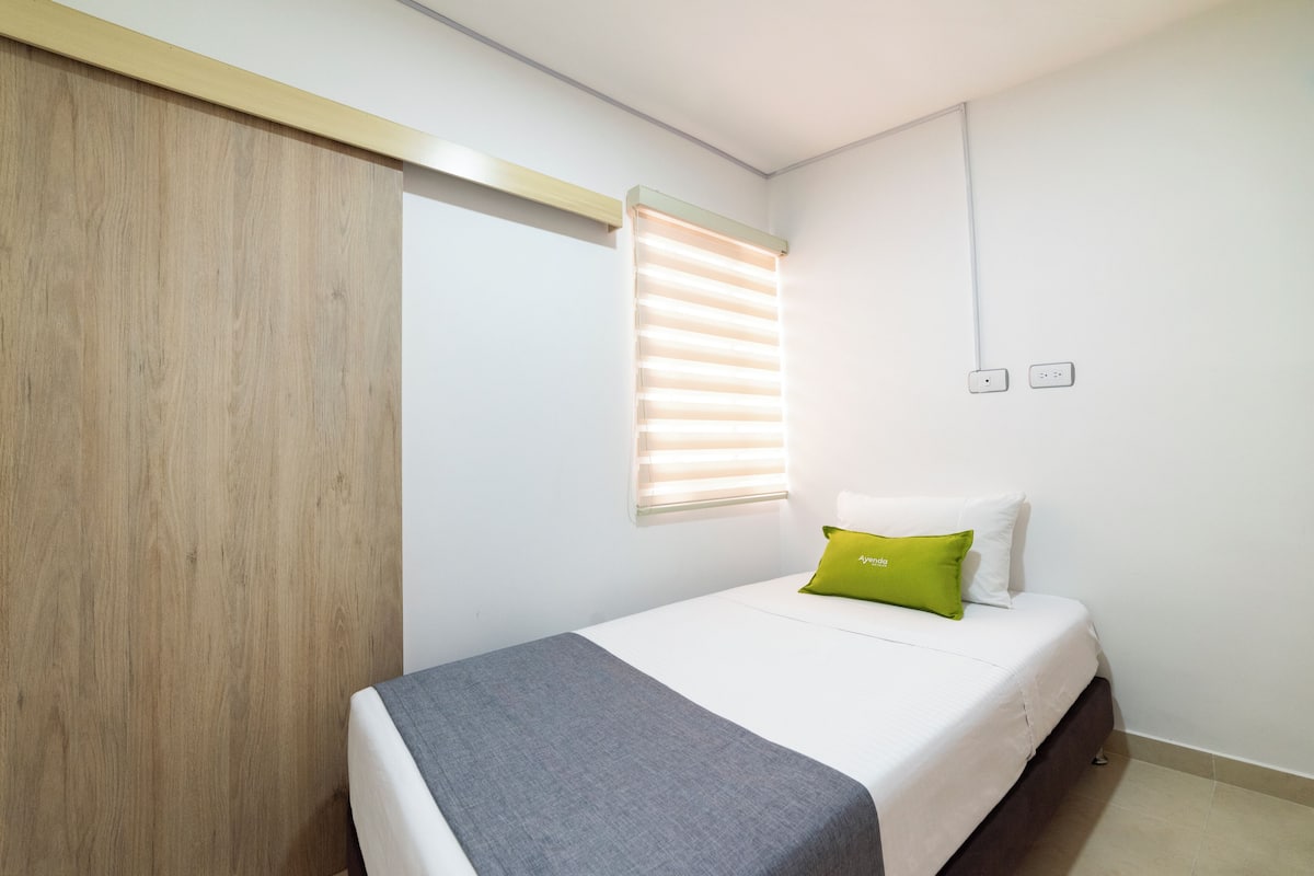 A bedroom with a bed, a window with blinds, a wooden wardrobe, and a bedside table.