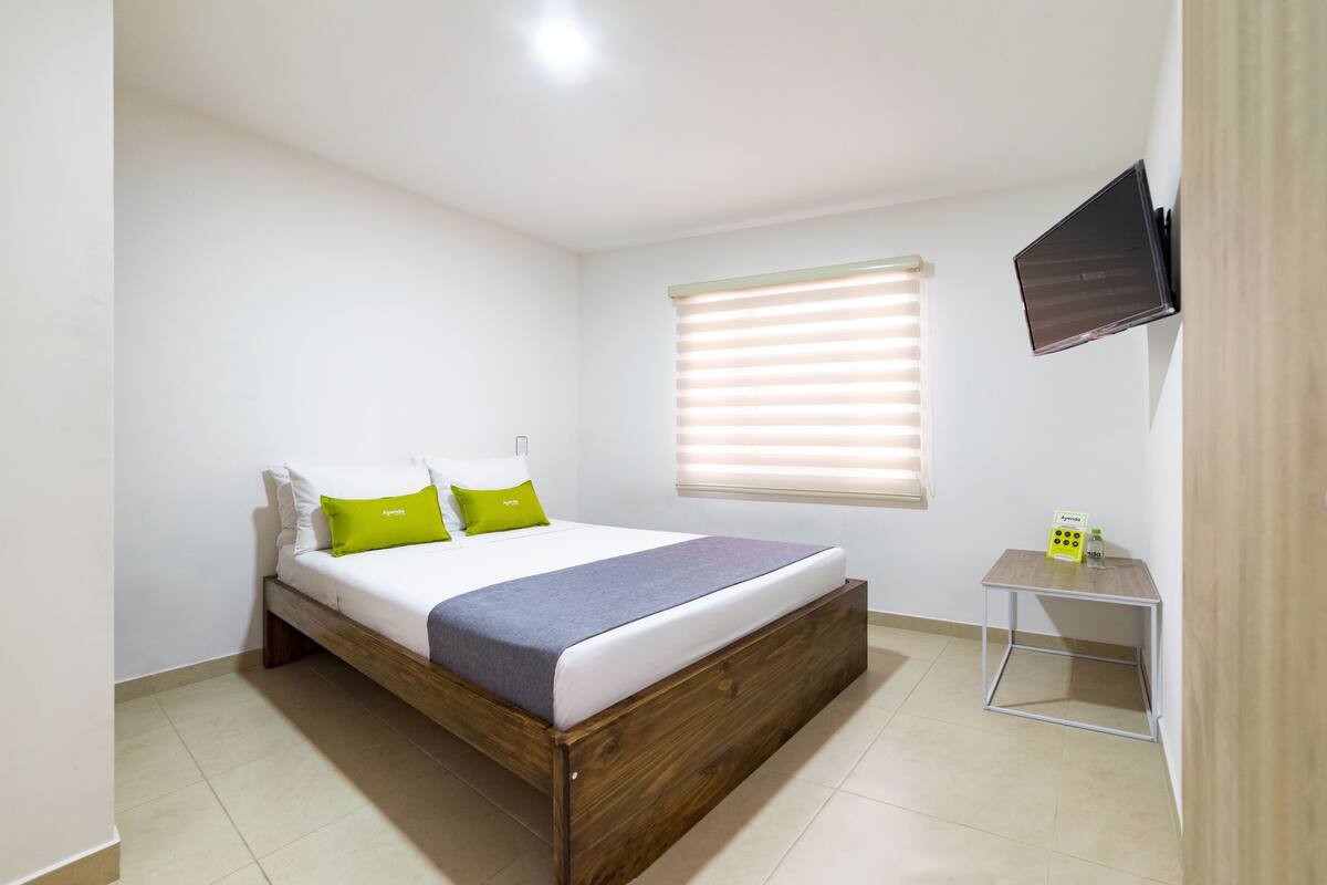 A bedroom with a wooden bed, white walls, a window with blinds, a small bedside table, and a flat-screen TV mounted on the wall.