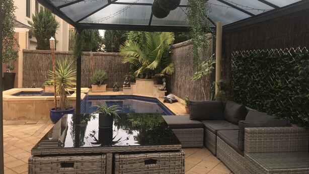 Ett takförsett utomhusområde med en pool, möbler av rotting och olika sorters krukväxter.