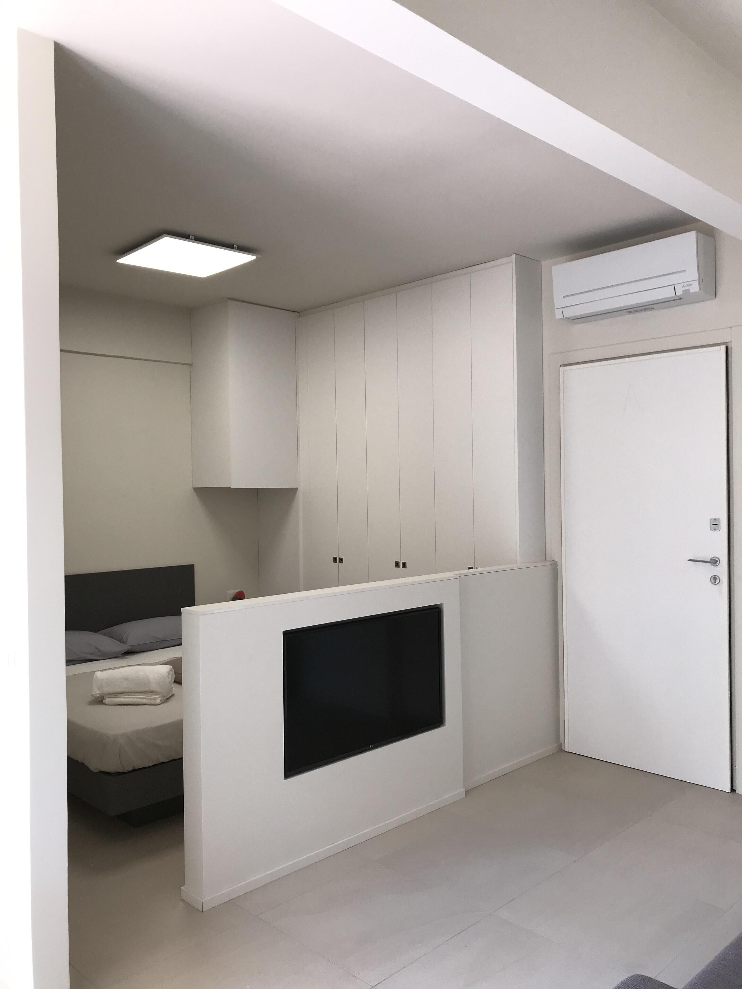 A modern room with a bed, built-in storage, a flat-screen TV, and an air conditioning unit.