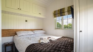 A bedroom with a wooden headboard, a large bed with a patterned bedspread, two rolled towels, a nightstand, and a window with a view of trees.