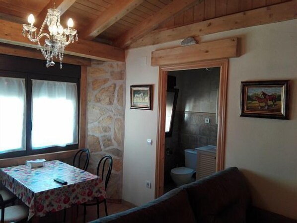 Ein gemütlicher Raum mit Holzbalkendecke, einer Chandelier, einem Esstisch mit Stühlen, einer Couch und einer Steinwand.