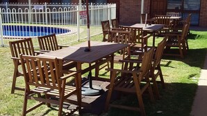 Mesas e cadeiras de madeira ao ar livre, com guarda-sóis, área da piscina cercada e um edifício com fachada de tijolos.