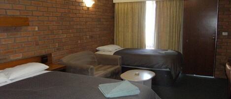 Habitación de hotel con pared de ladrillo, una cama grande, dos camas más pequeñas, una mesa redonda y una puerta.