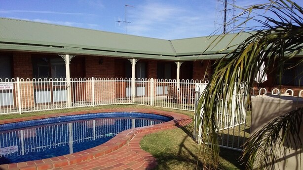 Uma piscina com borda de tijolos, cercada por uma cerca branca, e um edifício com telhado verde ao fundo.