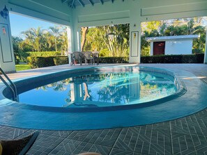 Una piscina con borde de baldosas, rodeada por una zona cubierta con sillas y una mesa, y vistas a la vegetación tropical.