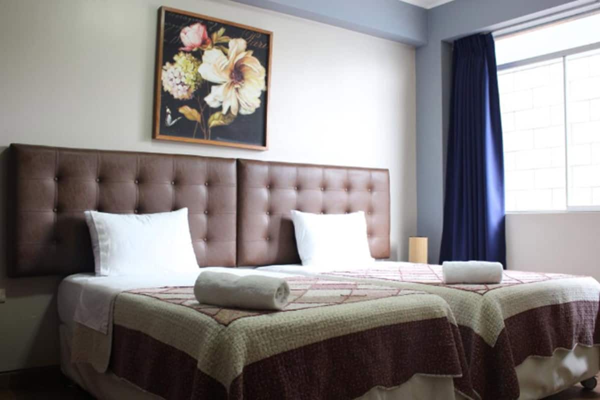 Uma cama de casal com cabeceira marrom, um quadro floral acima e uma janela com cortinas azuis.