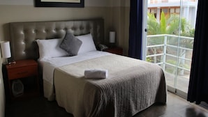 A hotel room with a large bed, a nightstand with a lamp, a telephone, and a window with a view of a building and greenery. - Gran Hotel Villa del Sur (San Vicente de Cañete)