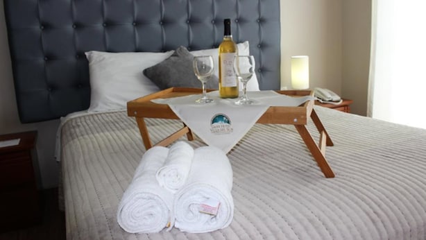 A neatly made bed with a grey headboard, a bedside table with a lamp, and a tray with a bottle of wine, glasses, and towels. - Gran Hotel Villa del Sur (San Vicente de Cañete)