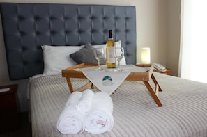 A neatly made bed with a grey headboard, a bedside table with a lamp, and a tray with a bottle of wine, glasses, and towels. - Gran Hotel Villa del Sur (San Vicente de Cañete)