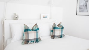 Una cama bien hecha con ropa blanca, dos almohadones escoceses, una manzana decorativa pequeña y un cuadro en la cabecera.