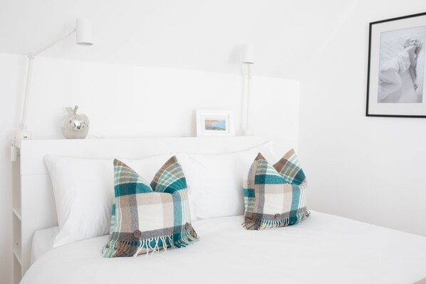 Un lit bien fait, avec une literie blanche, deux coussins à carreaux, une petite pomme décorative et un tableau encadré accroché au chevet.