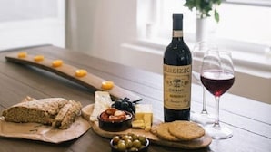 Una mesa de madera con una botella de Chianti, una copa de vino tinto, queso, aceitunas, galletas y pan.