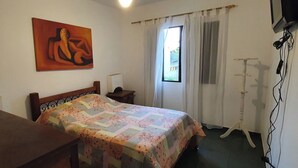 Un dormitorio con cama, escritorio, silla, un cuadro en la pared y ventana con cortinas.
