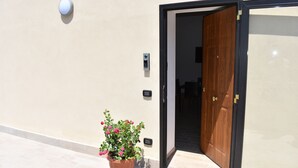 Une entrée moderne avec une porte en bois, un pot de fleurs orné de fleurs roses et un système d’interphone fixé au mur.