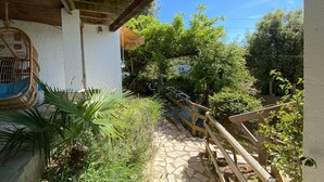 A stone pathway leading to a covered area with a wooden railing, surrounded by lush greenery and a clear blue sky. - Beautiful studio/apartment with air conditioning and use of pool.  (Begur)