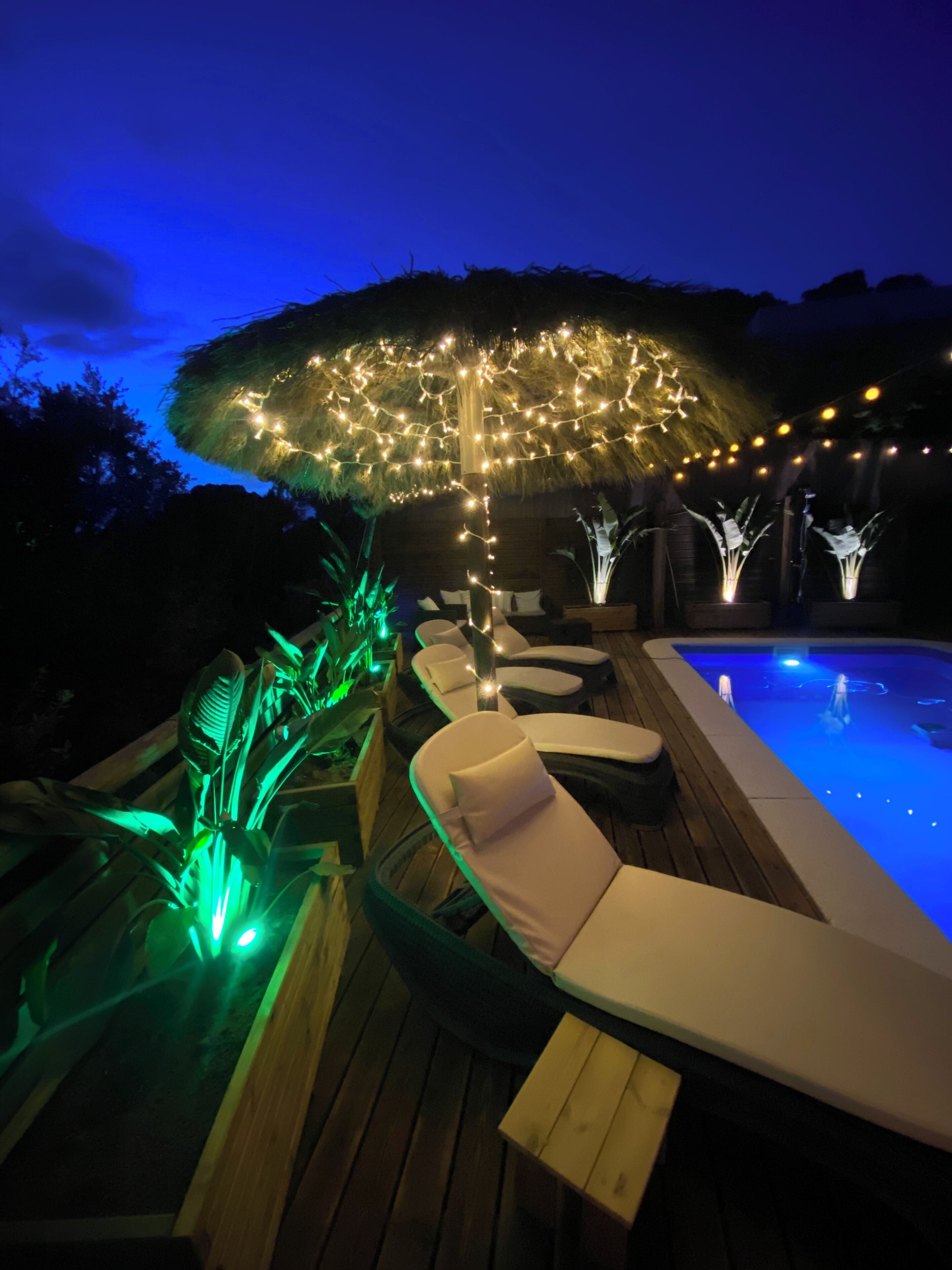 Zona de piscina con sillones reclinables, una sombrilla con luces de cadena y un elemento acuático.